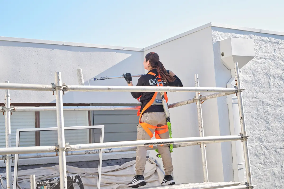 Professional painter standing on scaffolding applying white paint to a building’s exterior wall using a roller, wearing safety harness and protective gear under clear daylight.