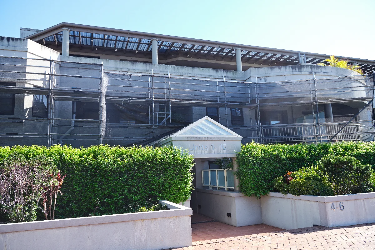 Building under renovation with scaffolding and netting along the facade, and a hedge-lined entrance.