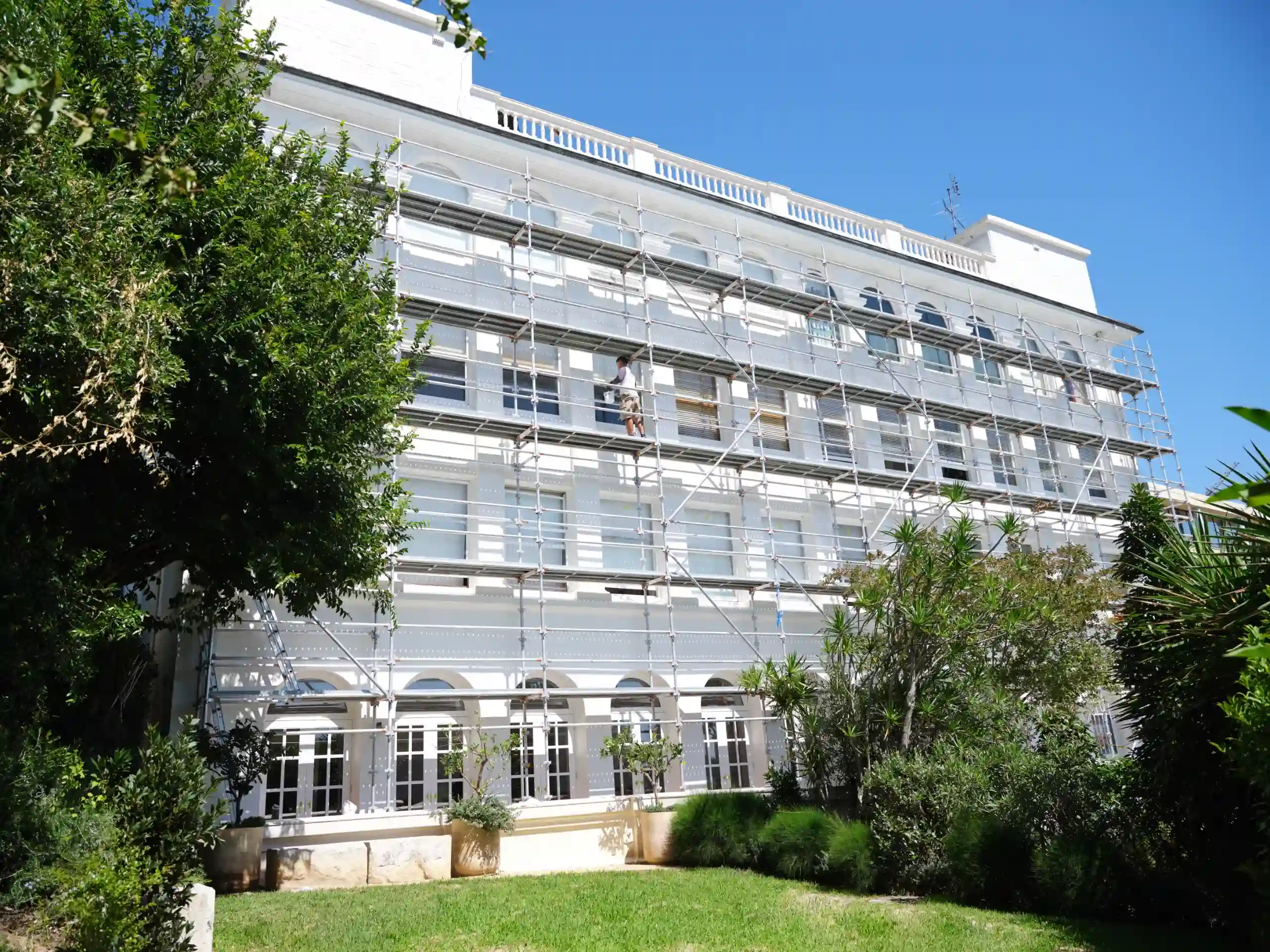 White multi-story building wrapped in metal scaffolding against a clear blue sky; green trees in the foreground.