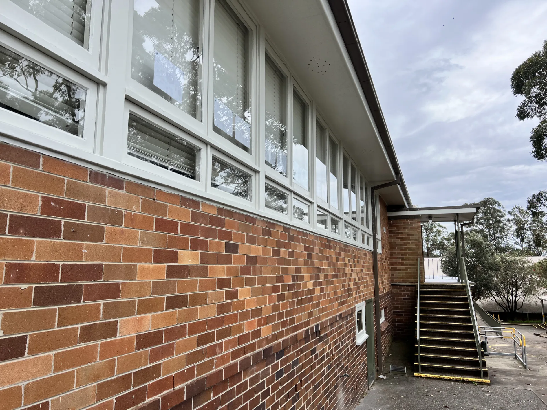 Exterior view of a red-brick school building with large white-framed windows and an outdoor staircase.